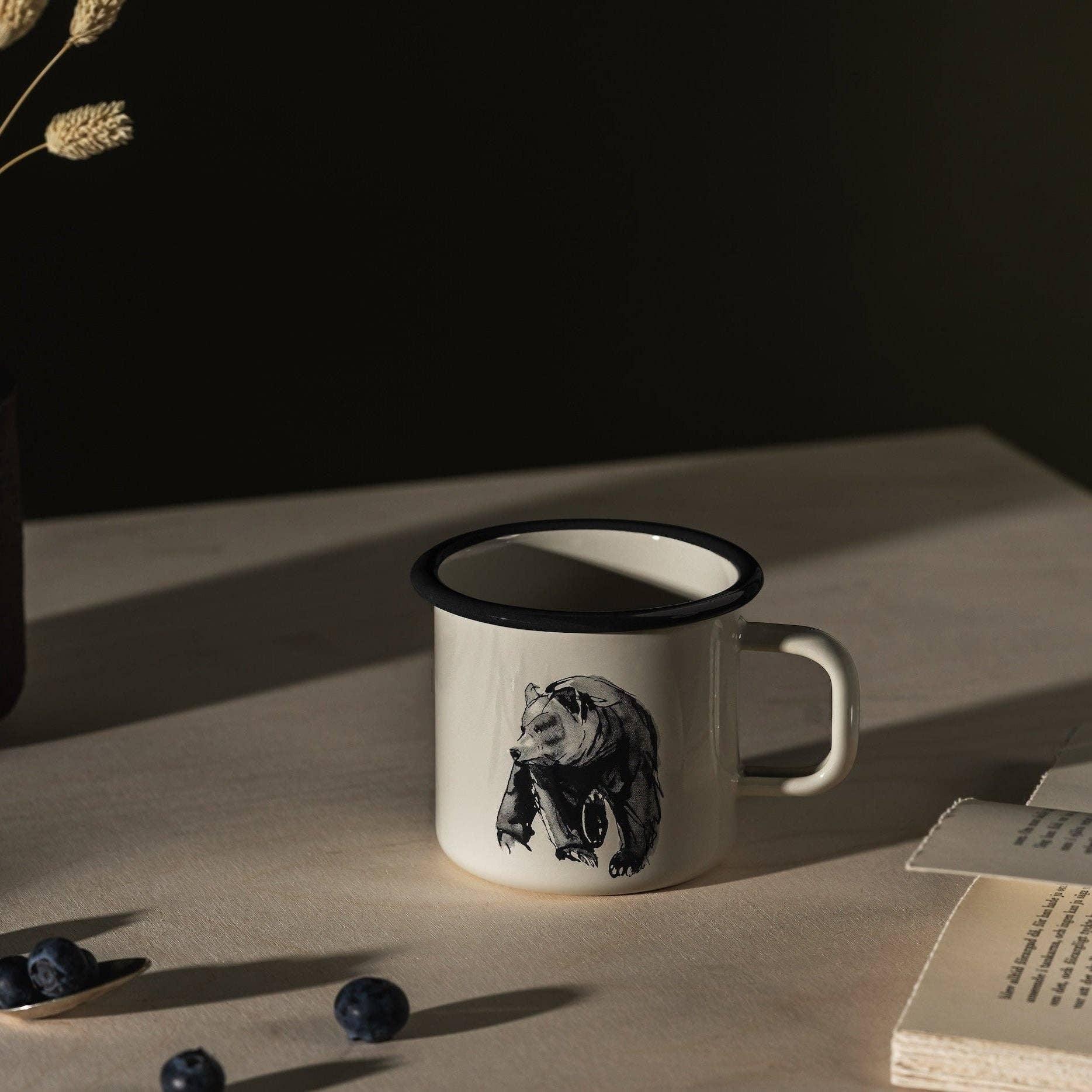 Wildo enamel mug with black bear print, cream color, on light wood table with blueberries