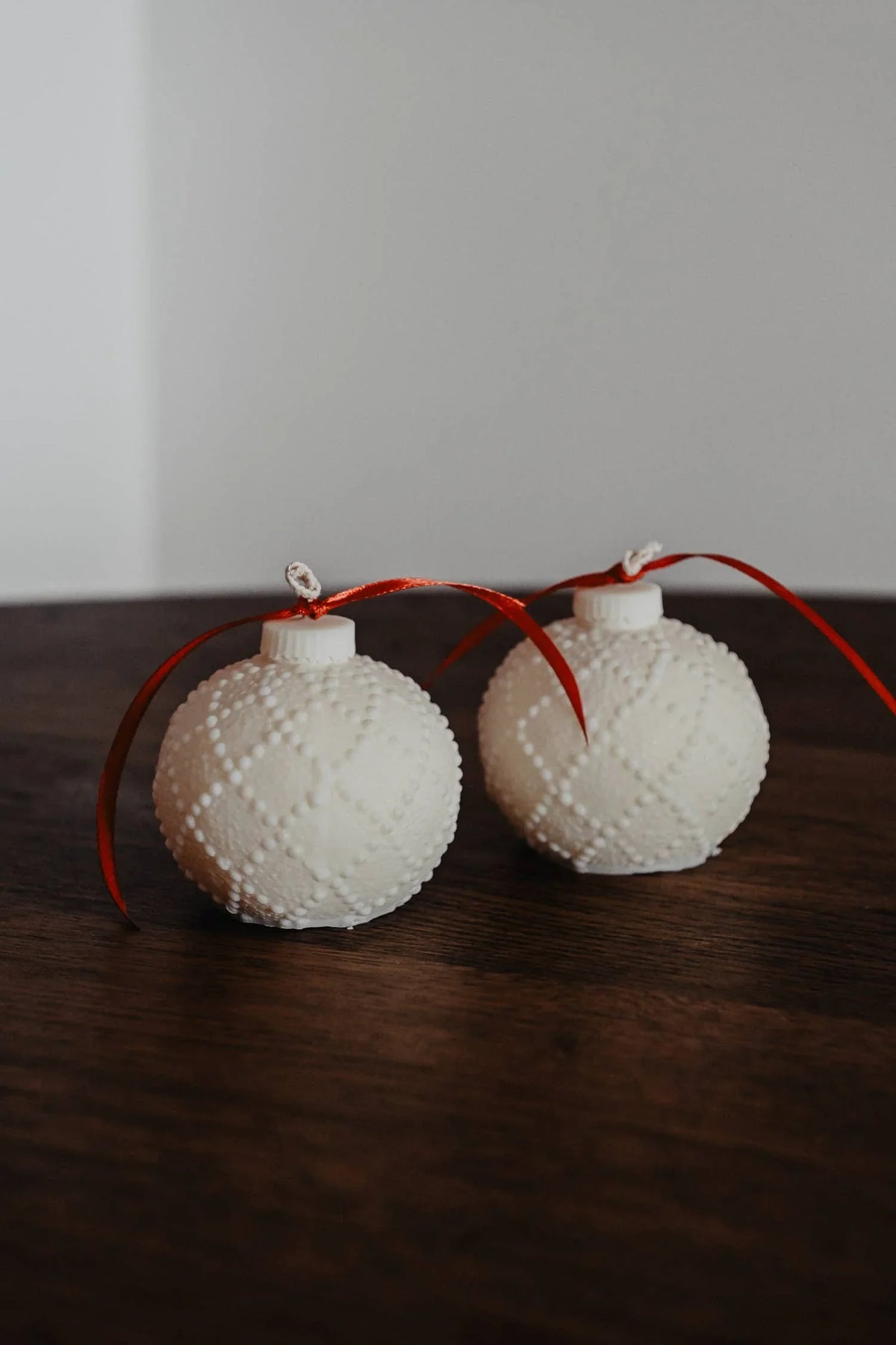 Two Studio Twocan white ceramic Christmas ornaments with red ribbons on wood table