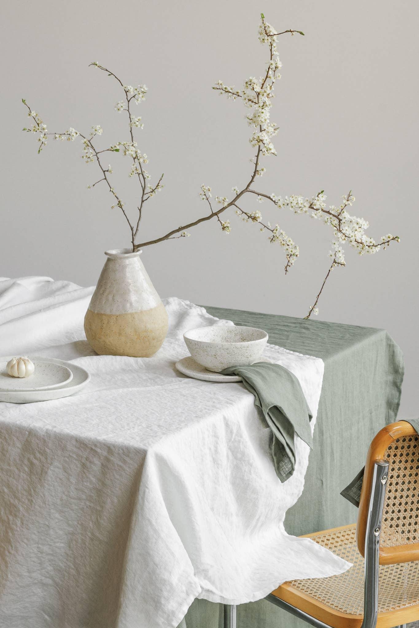 Pomponner stoneware vase with white and beige finish, linen tablecloth, ceramic bowl, and branch decor on minimalist dining table.