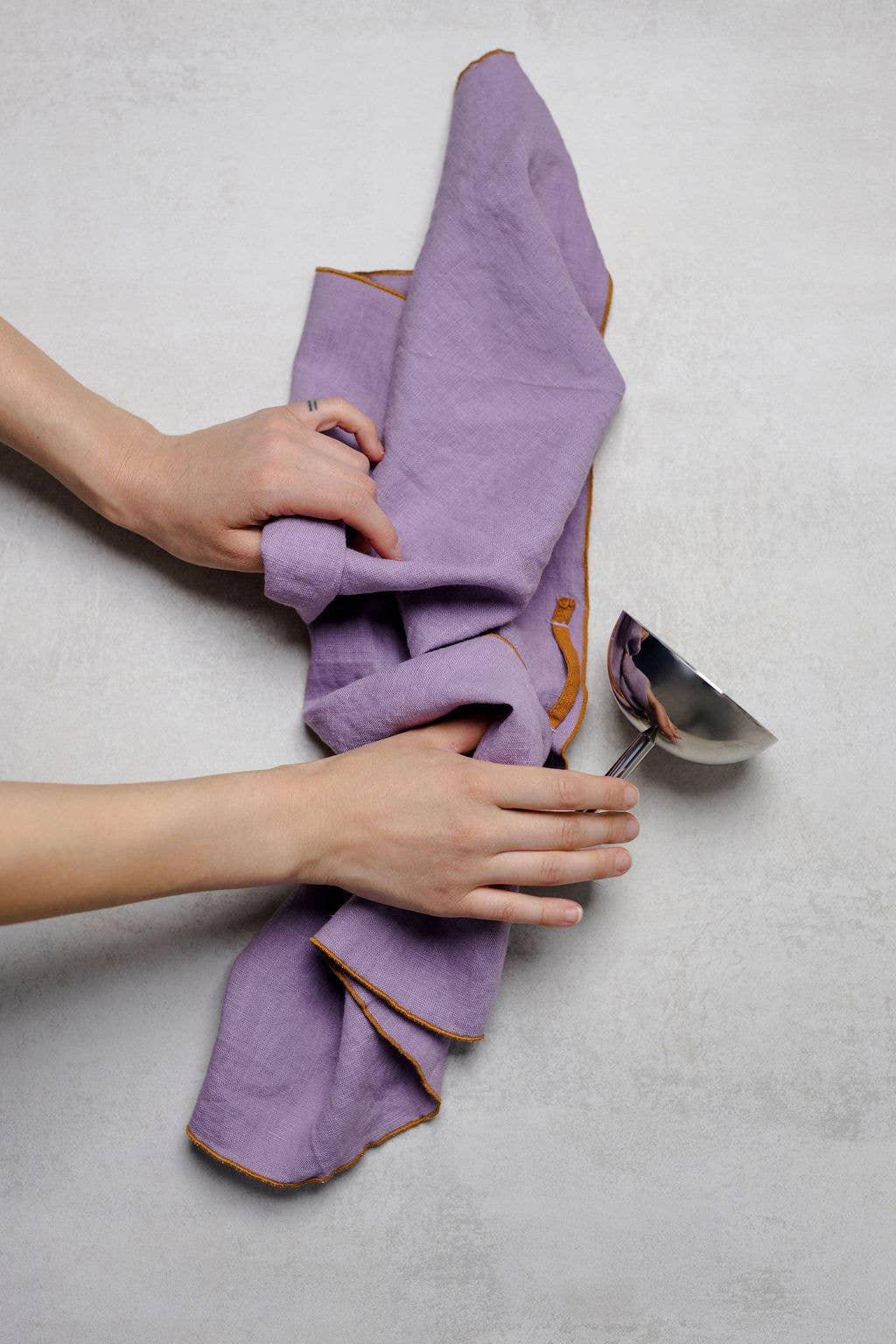 Linen Tales lavender linen napkin with orange trim held near a silver ladle on light surface