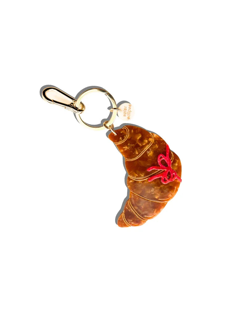 Sugar Eclipse croissant keychain, brown resin with gold hardware and red ribbon accent
