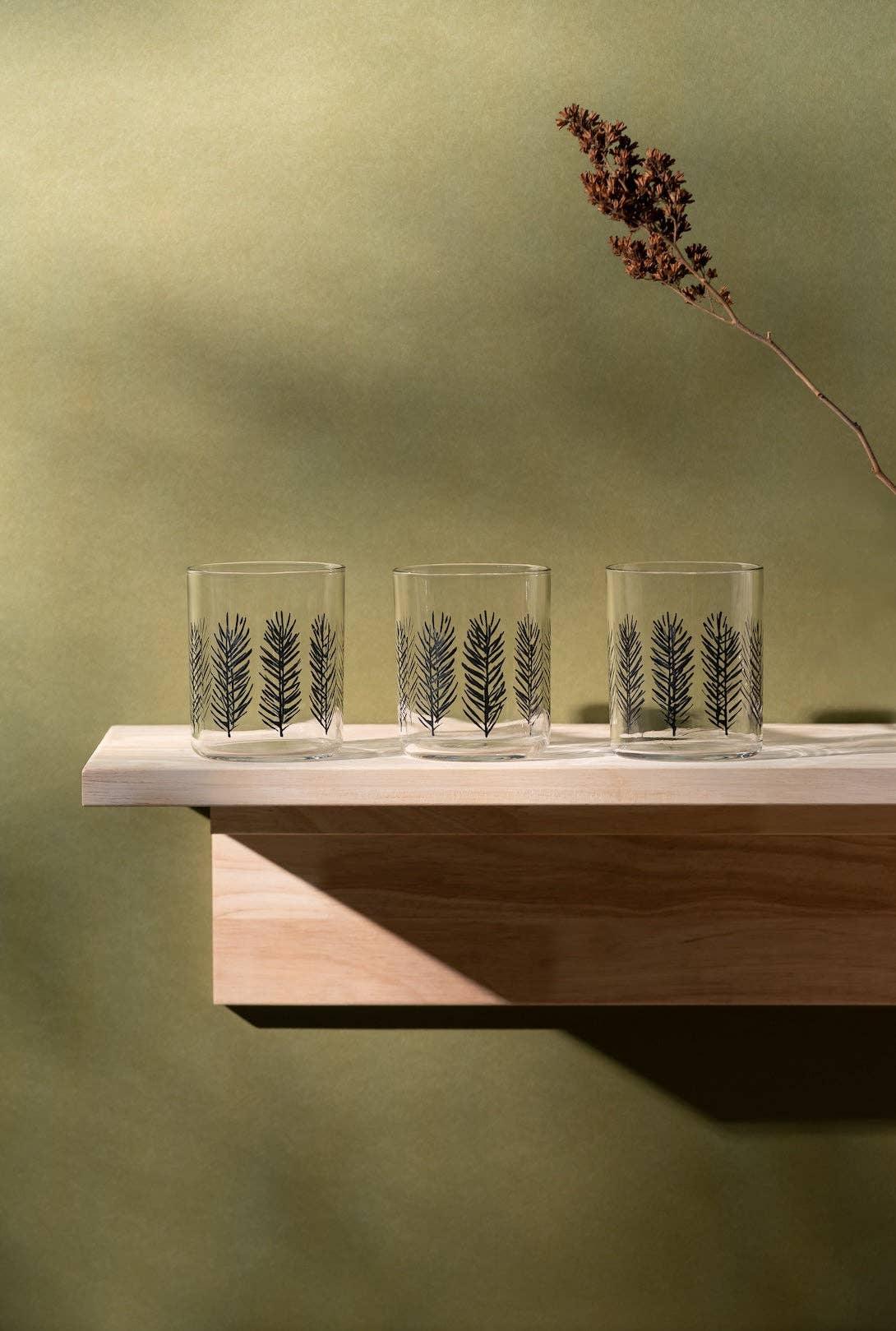Three Norden clear glass tumblers with black leaf pattern on a light wood shelf, green background