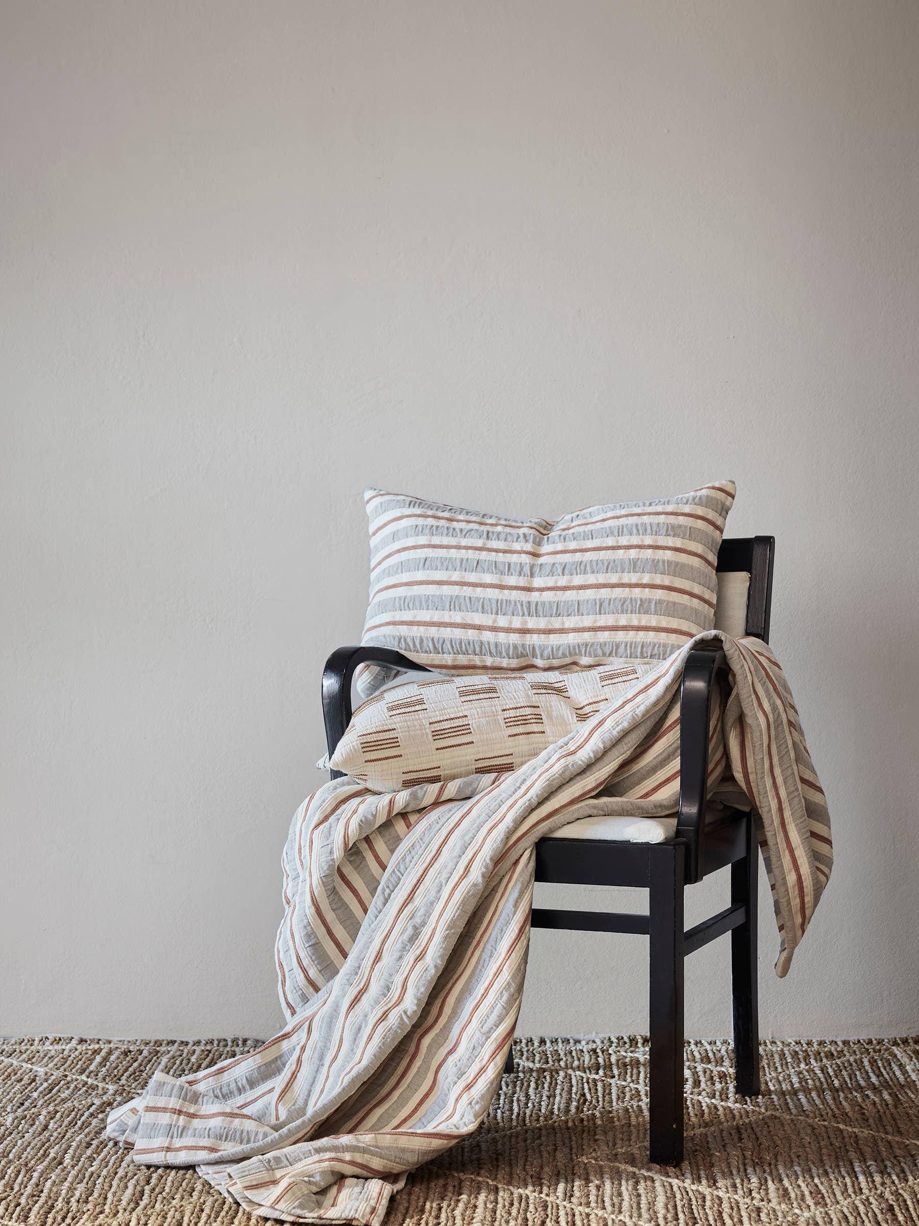 Tensira striped cotton throw blanket and pillow in beige, brown, and grey on black chair