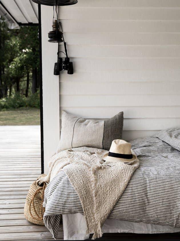 Linum striped linen bedding with beige pillow and throw on outdoor bed, neutral straw accessories.