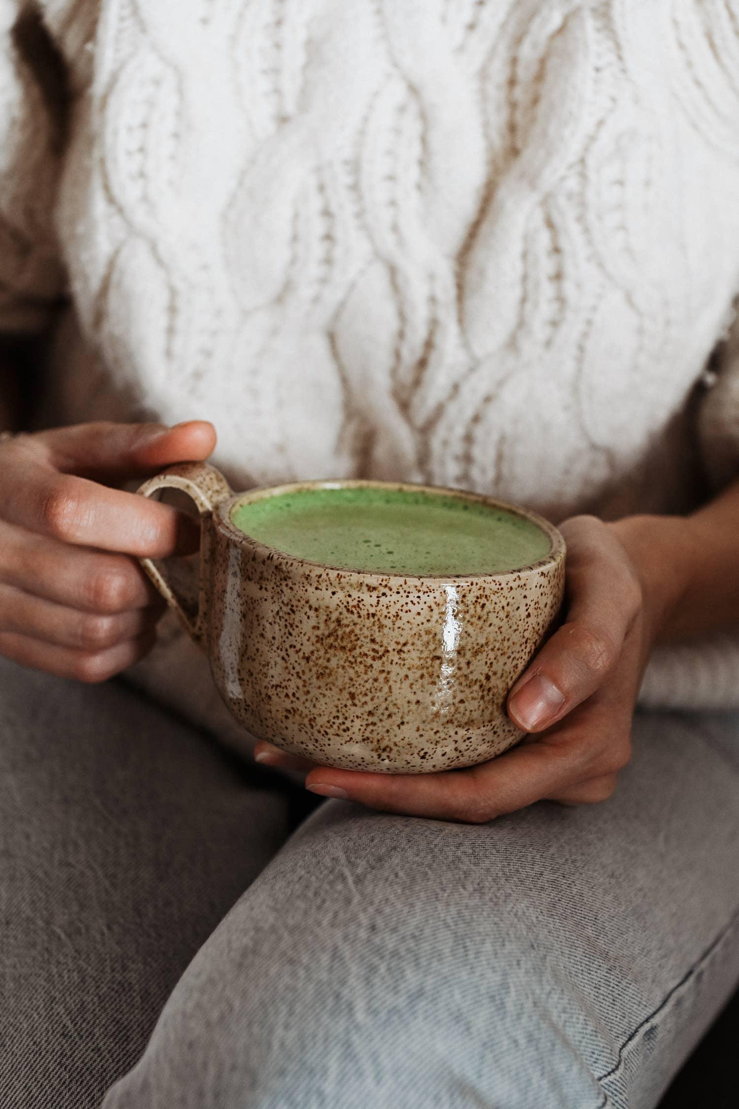 Hand holding Speckled ceramic mug with green matcha latte, cozy knit sweater background