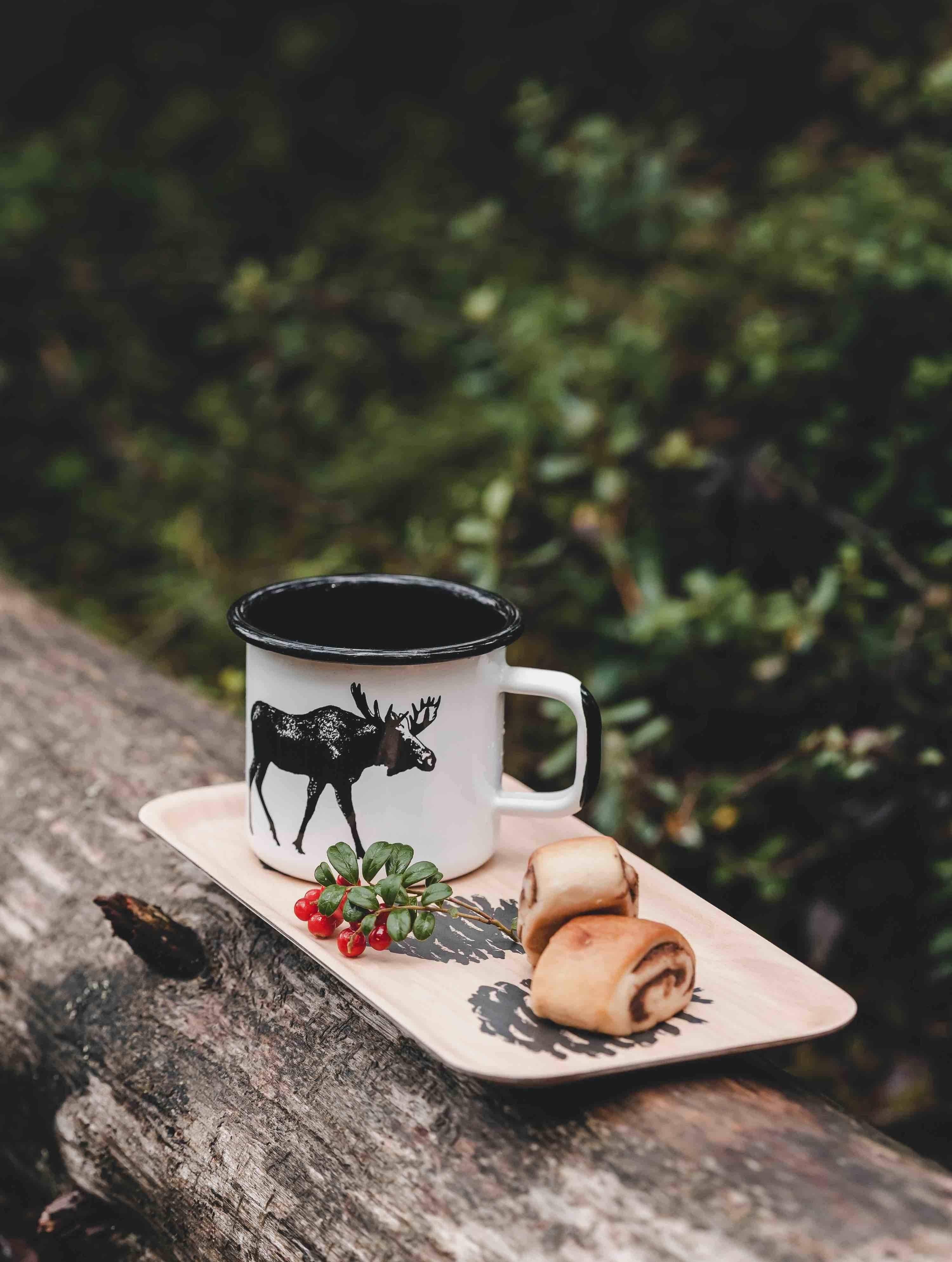 Muurla enamel mug with moose print, white and black, on wood tray with pastries outdoors