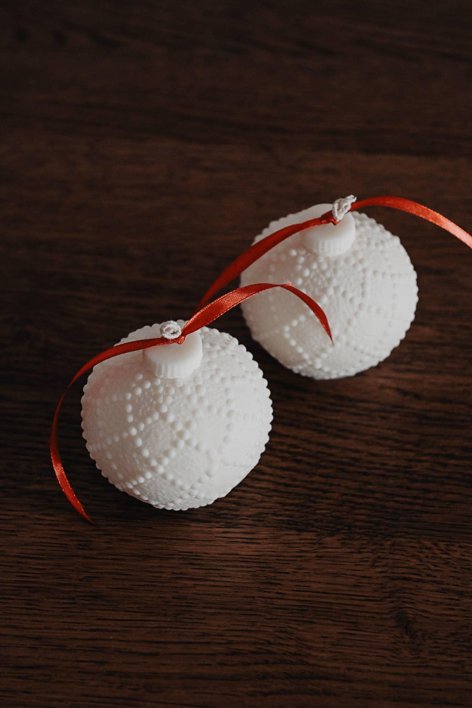 Two minimalist white ceramic Christmas ornaments with red ribbons on wood surface