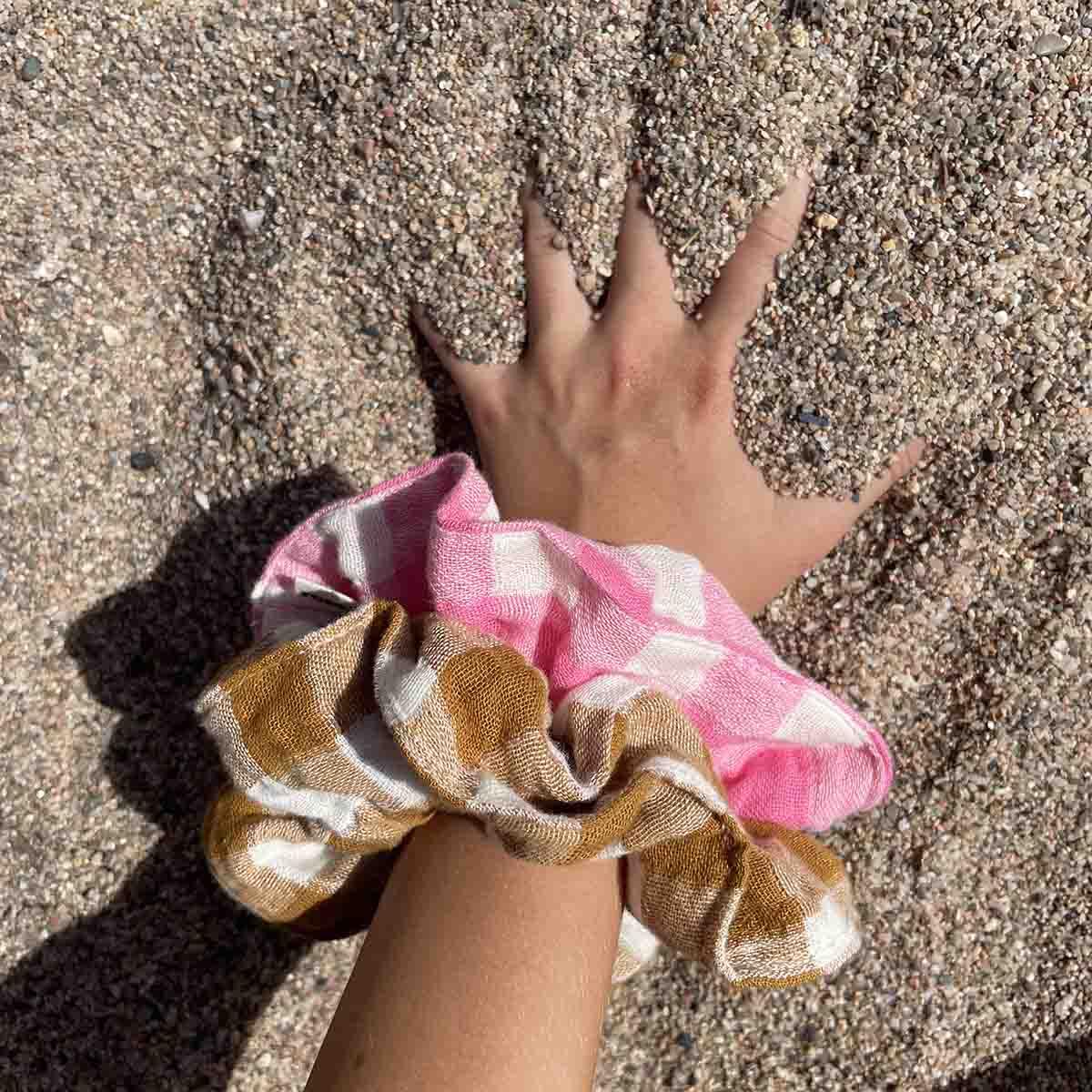 Ametrine Studio linen scrunchies in pink and brown gingham on wrist, beach sand background