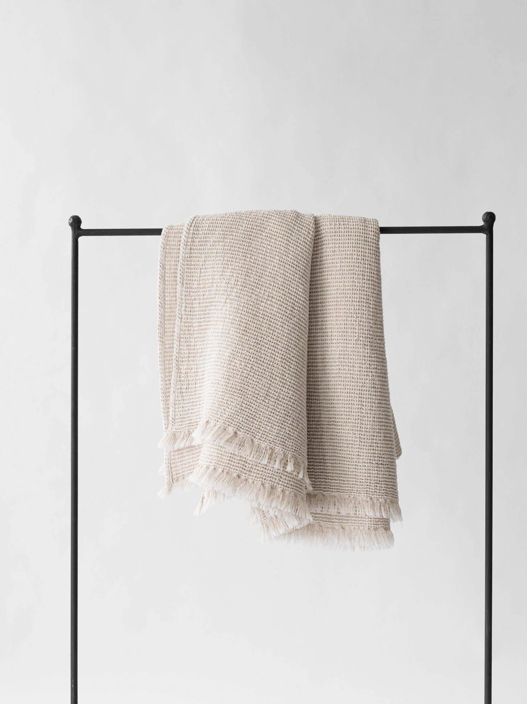 Tensira beige cotton throw blanket with fringe, displayed on black metal rack