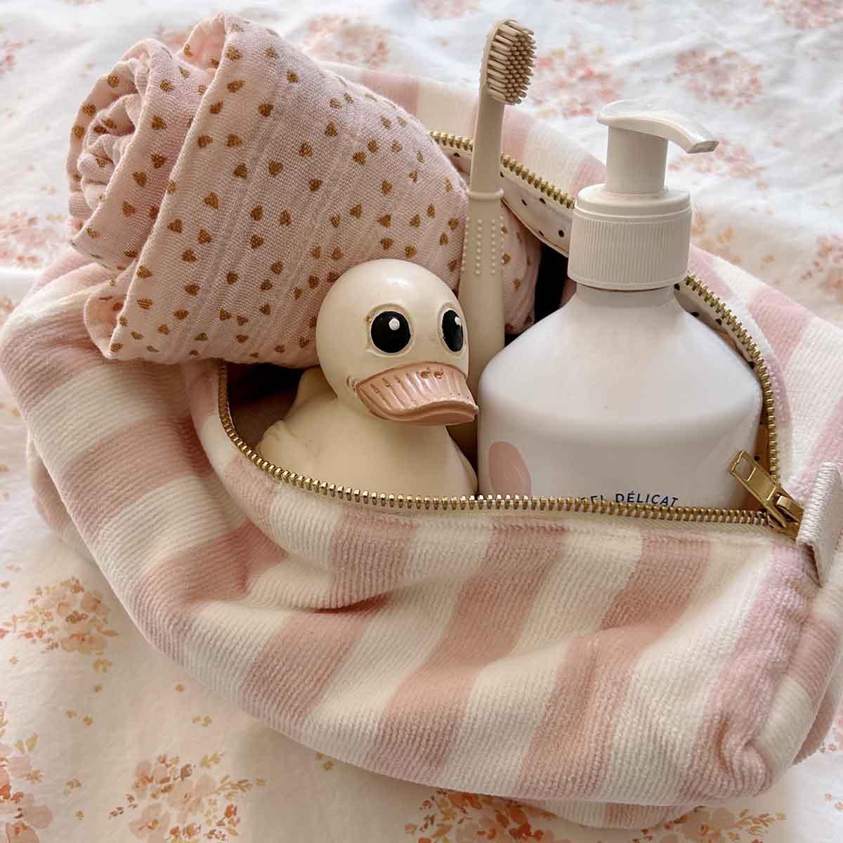 Mushie striped toiletry bag with pink baby towel, rubber duck, cream pump bottle, and toothbrush.
