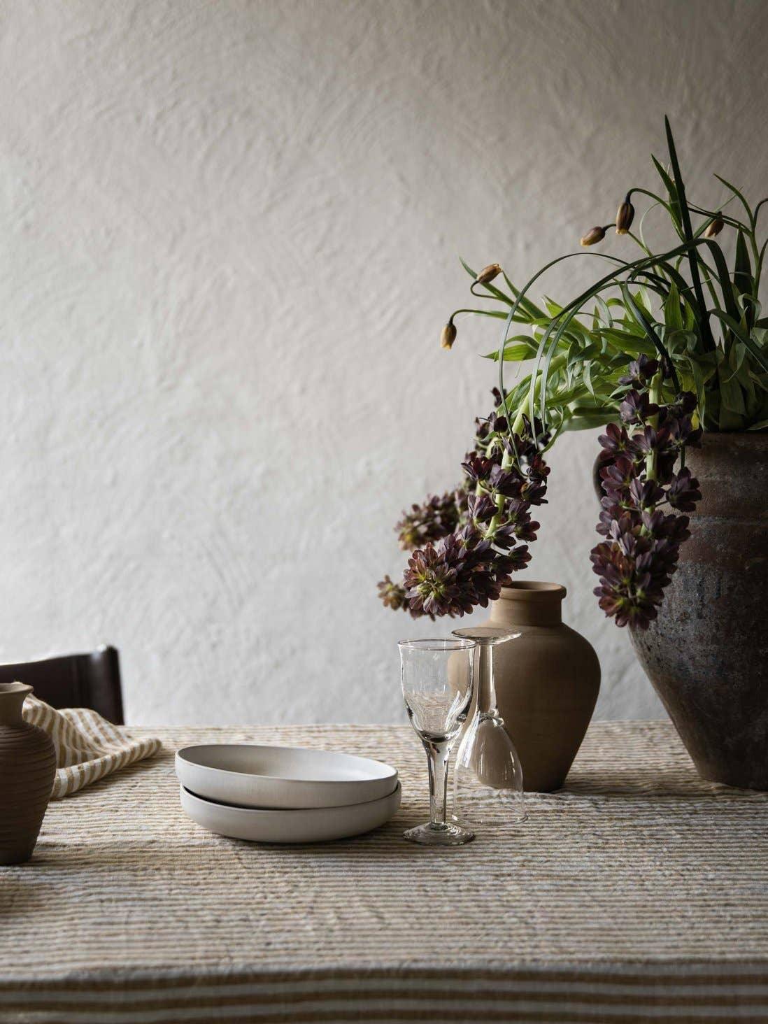 Hand-thrown ceramic bowls, glassware, earthy vases, and dark blooms on striped linen tablecloth