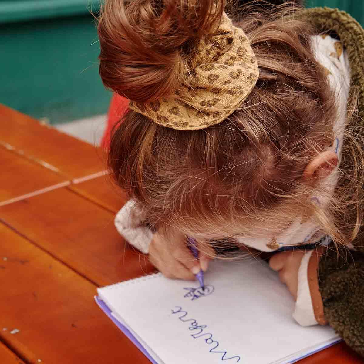 Child wearing leopard print scrunchie, drawing with blue pen on white paper at wooden table