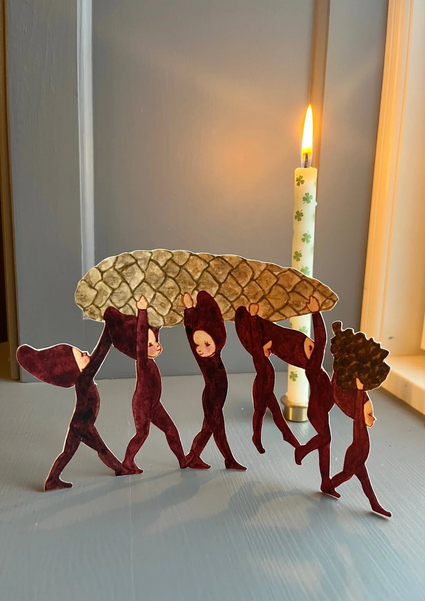 Waldorf-style Grimms paper figures carrying pinecone, burgundy and brown, candle in background