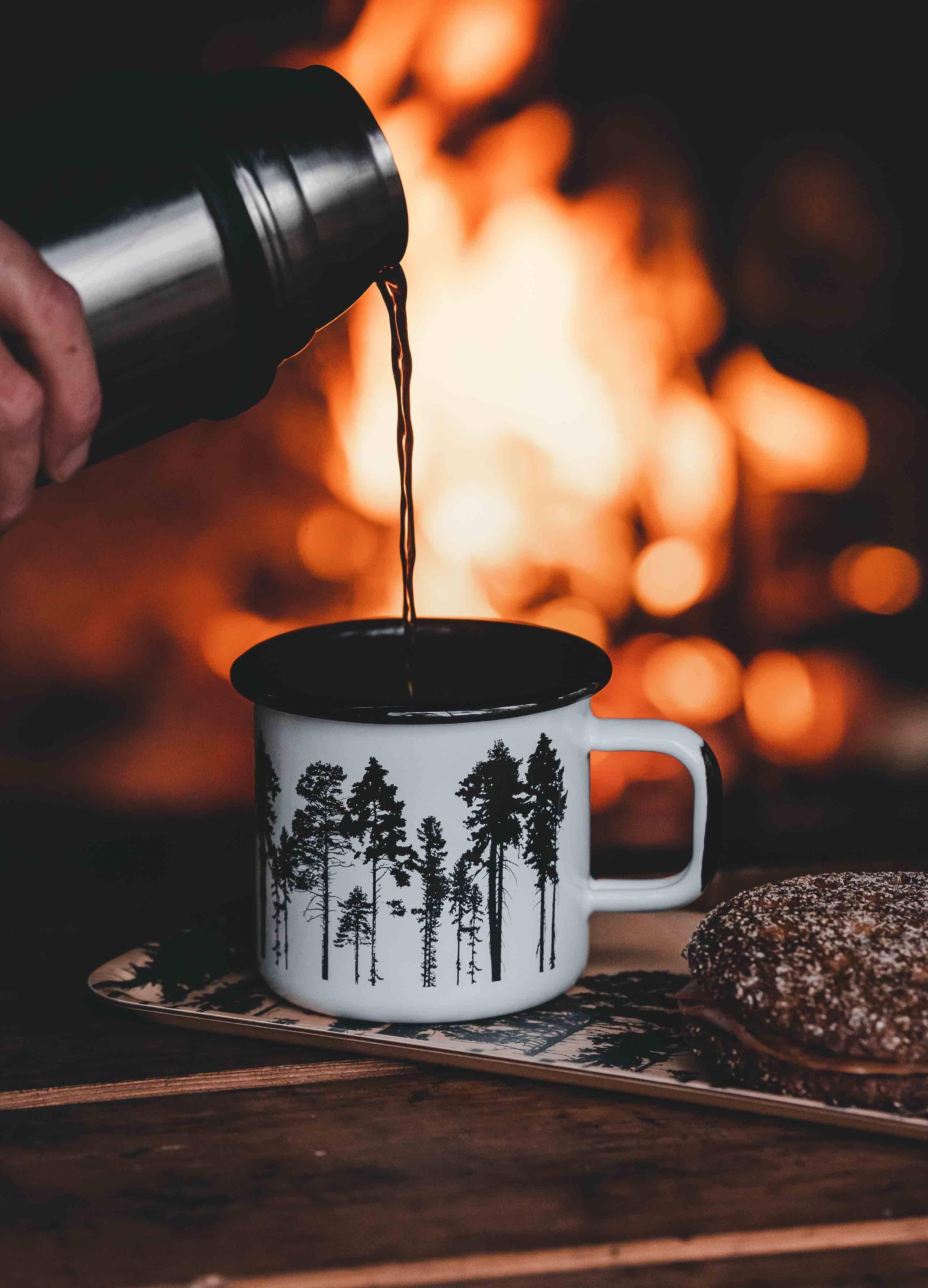 Enamel forest-print mug by Nordic Woods, white and black, coffee pour, fireplace background