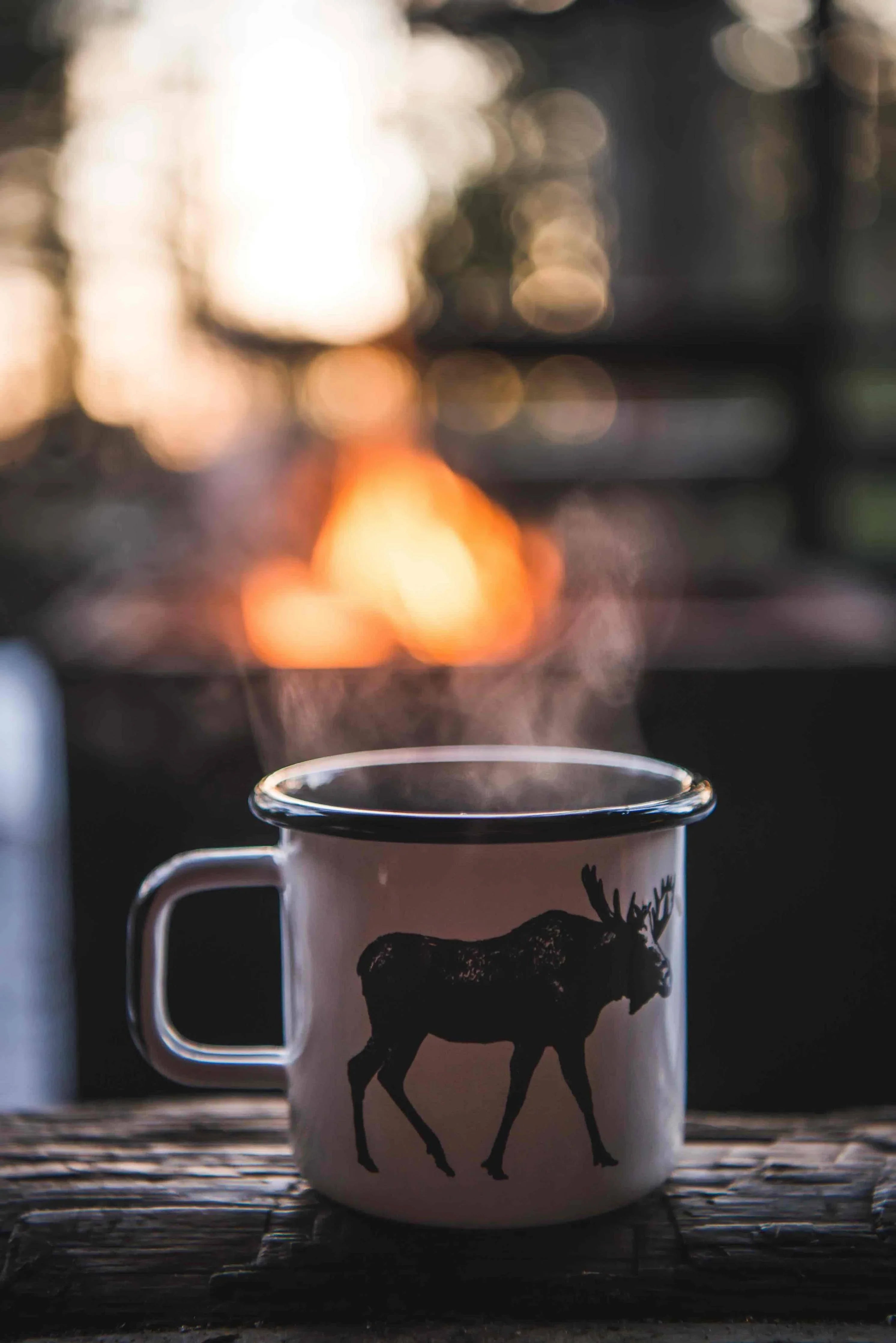 White enamel mug with moose graphic, steaming by campfire outdoors, rustic premium style.