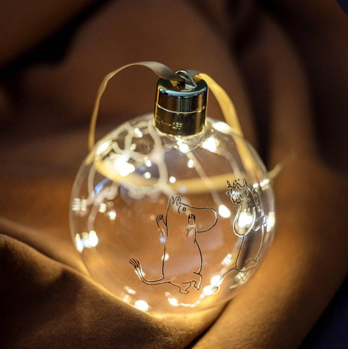 Moomin clear glass Christmas bauble with LED lights and gold ribbon on brown fabric