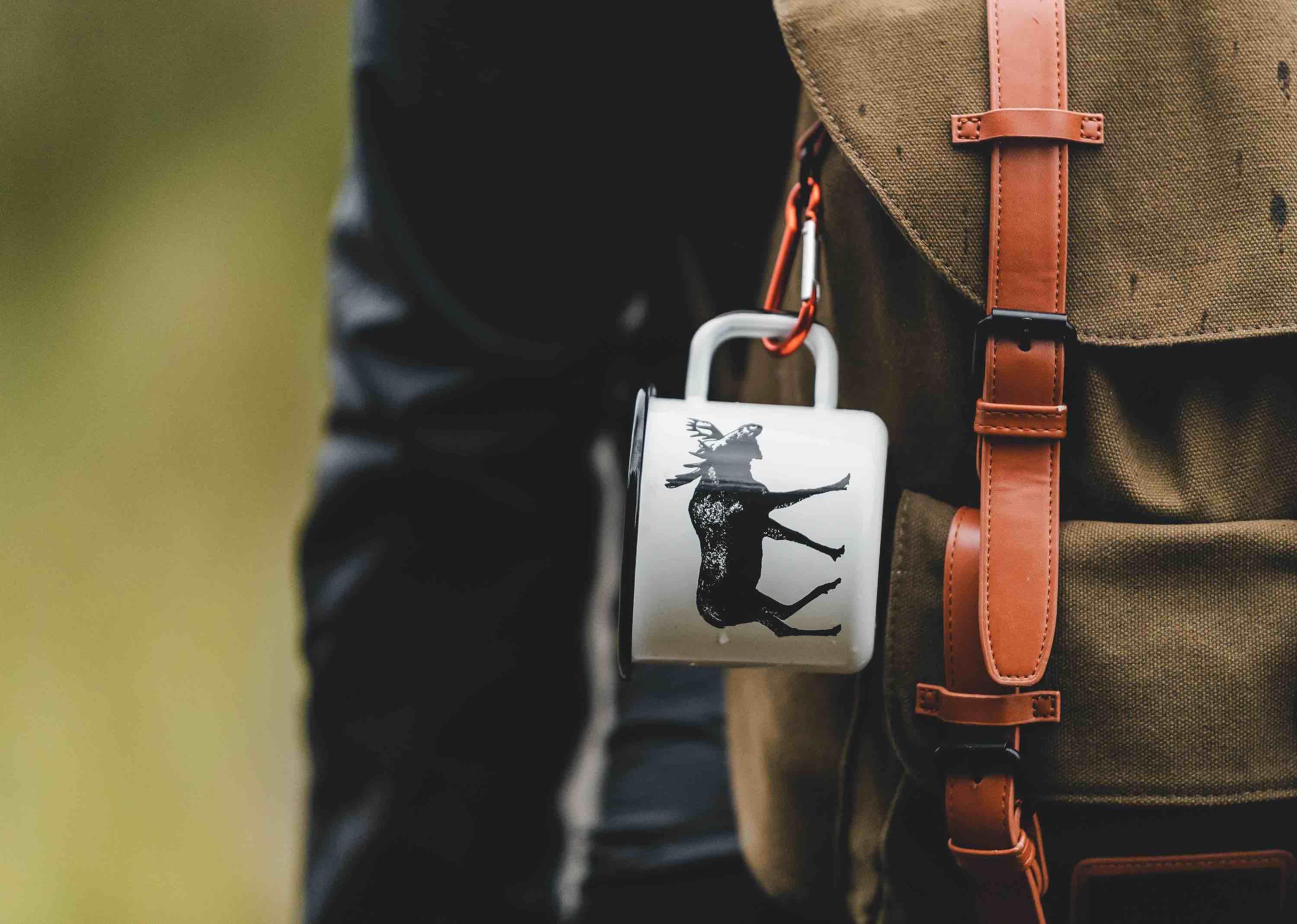 Moose graphic enamel mug on tan canvas backpack with leather straps outdoors