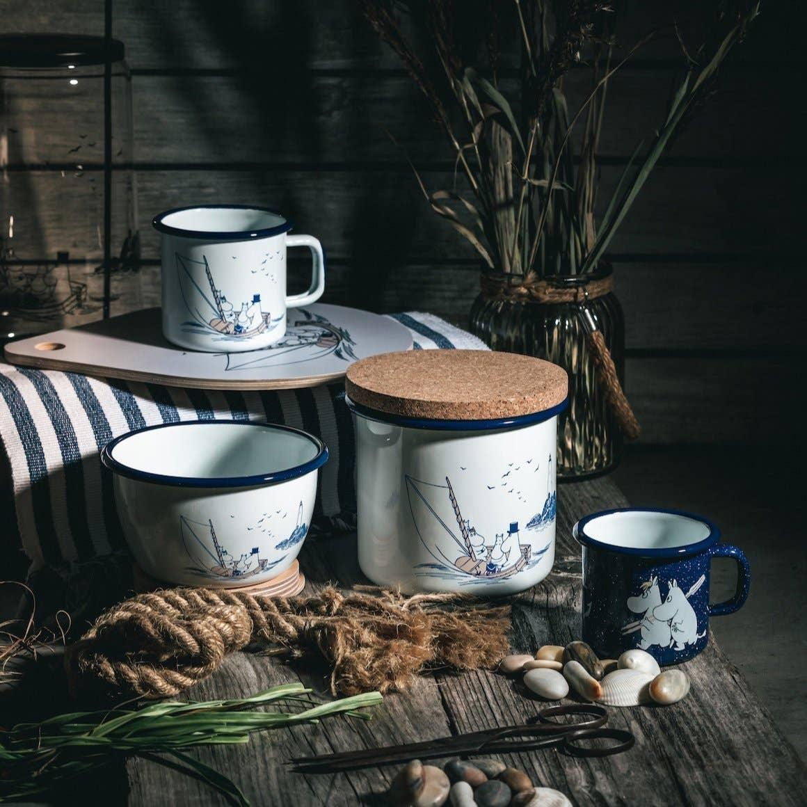 Moomin enamel mugs, bowl, and jar with cork lid, white and blue, on rustic wood table