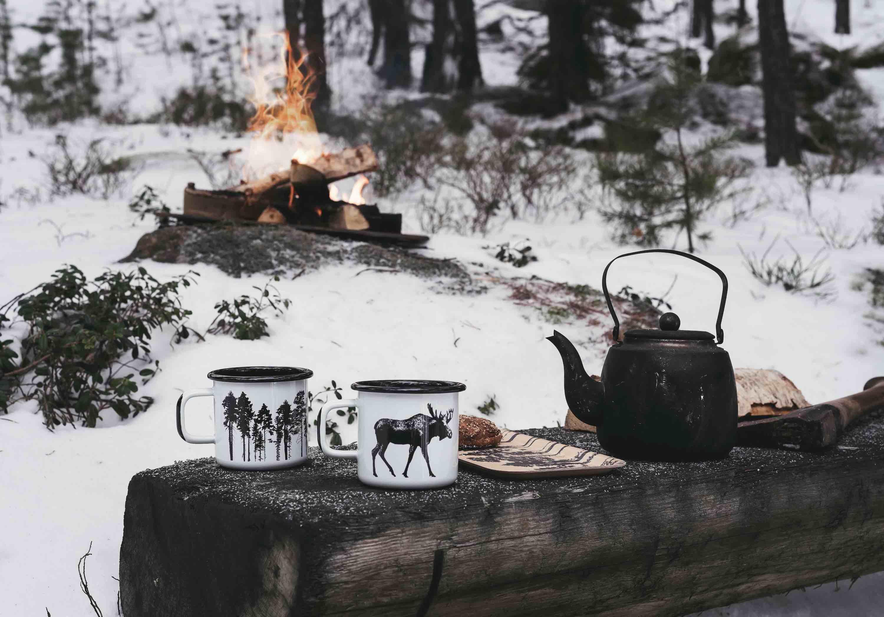 Nordhusen enamel outdoor mugs with woodland and moose graphics, black kettle on snowy log, campfire in background