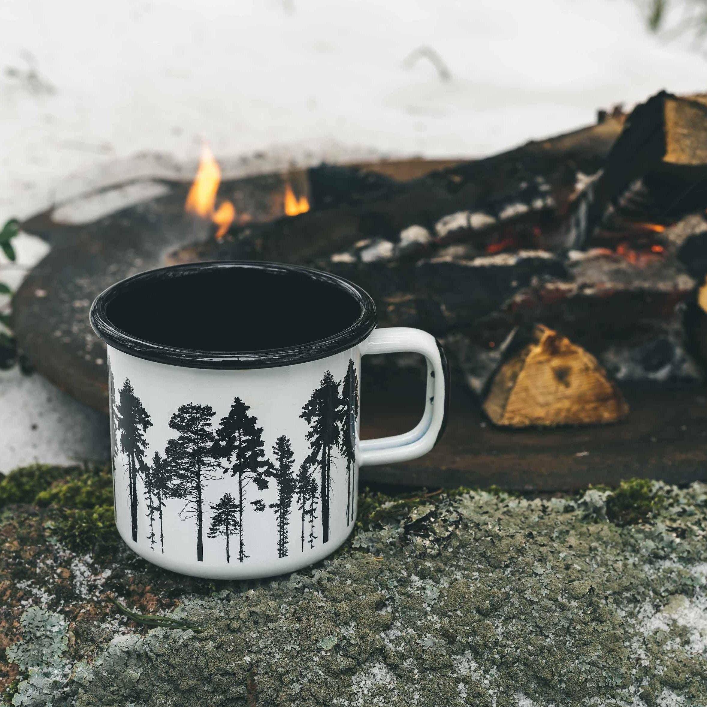 Muurla enamel mug with black tree design, outdoors on stone by campfire in winter