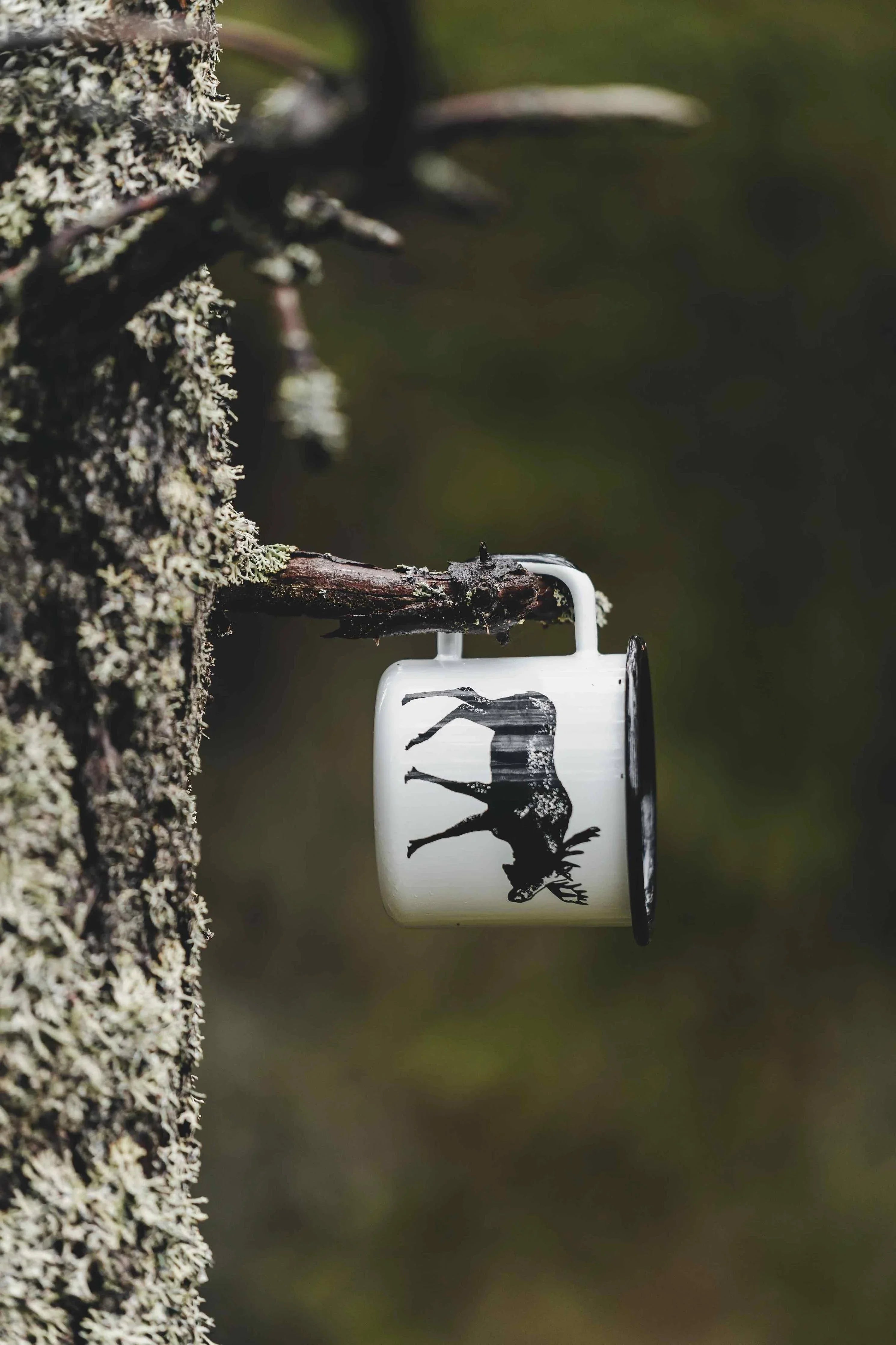 Nordic Moose white enamel mug with black rim and moose print hanging on tree branch outdoors