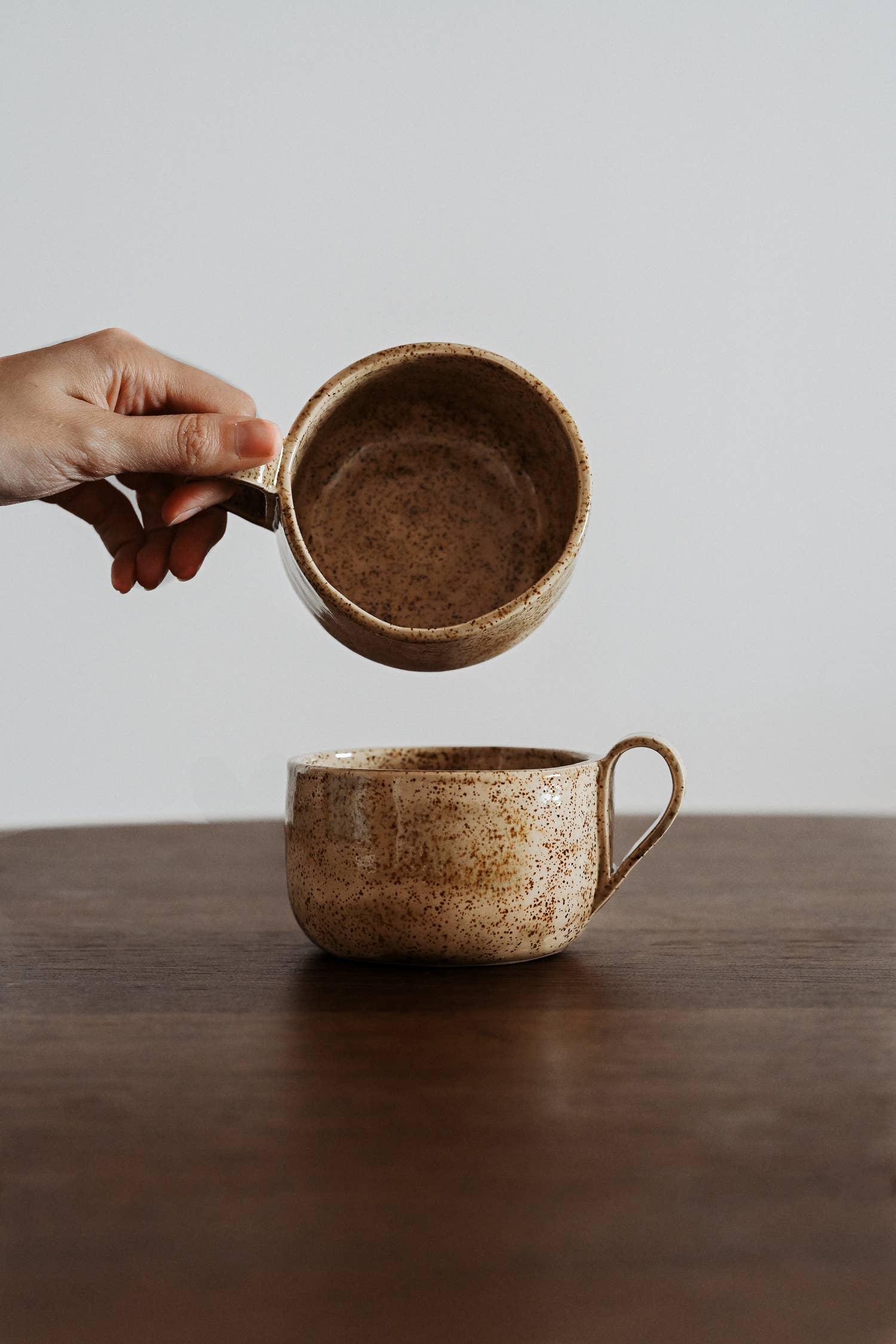 Hand holding Hasami porcelain speckled beige ceramic mug above matching cup on wood table