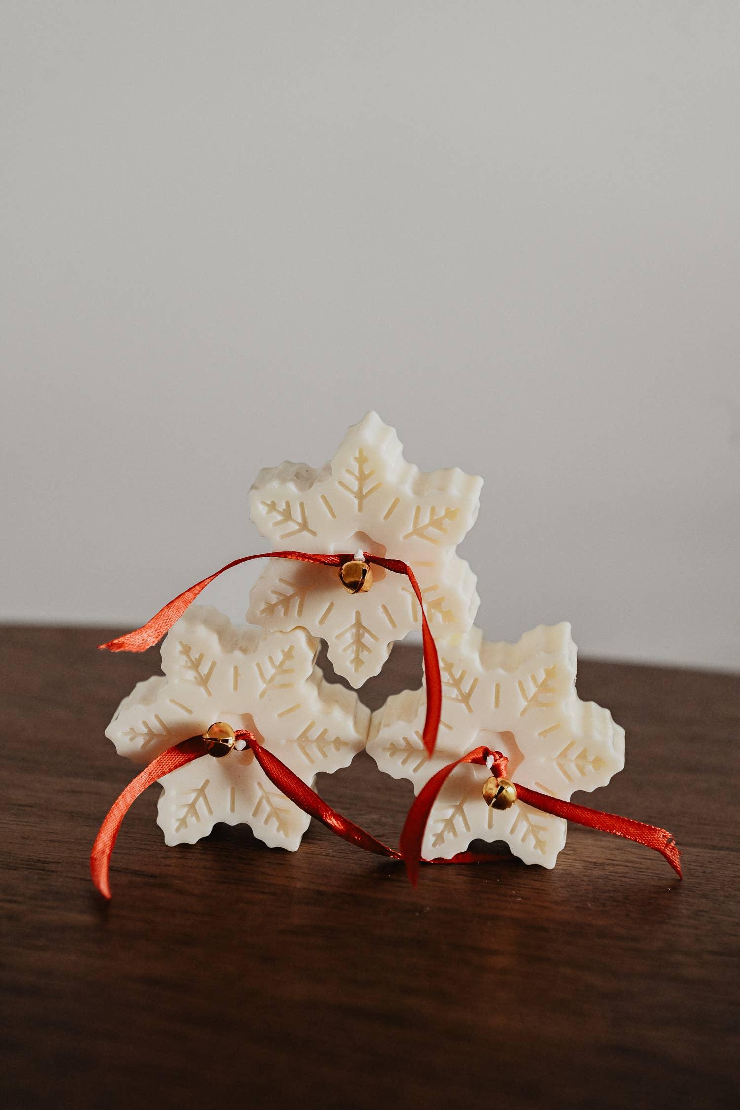 Snowflake-shaped white wax ornaments with red ribbon and gold bell, displayed on wood