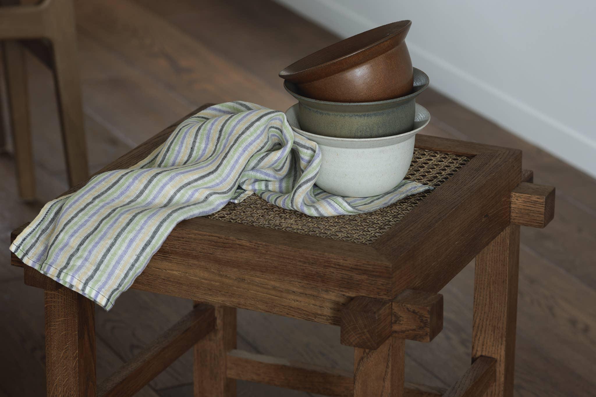 Stacked Hasami ceramic bowls in brown, grey, and white on a wooden woven stool with striped linen towel