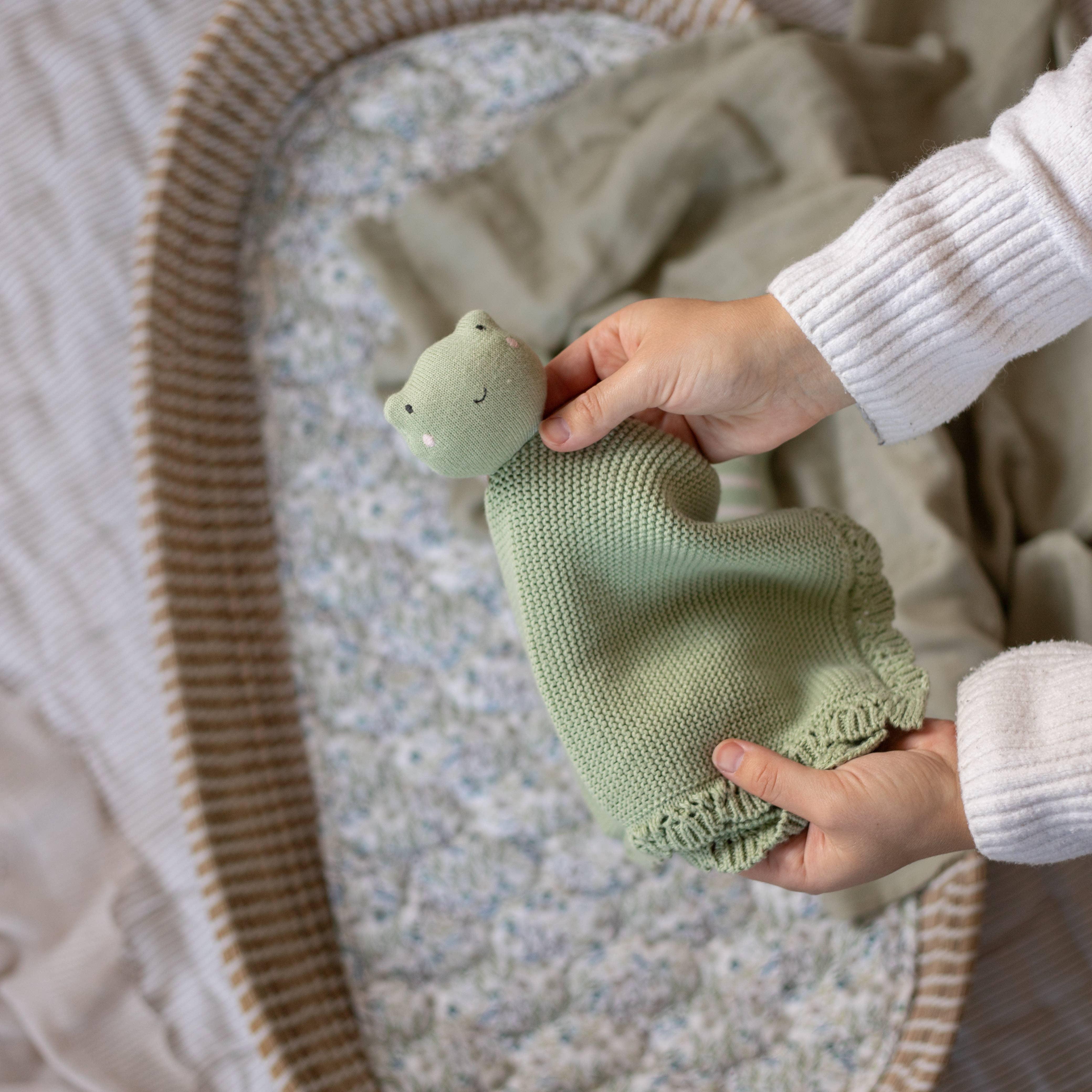 SAGA Copenhagen green knitted cotton frog comforter held over a neutral baby nest