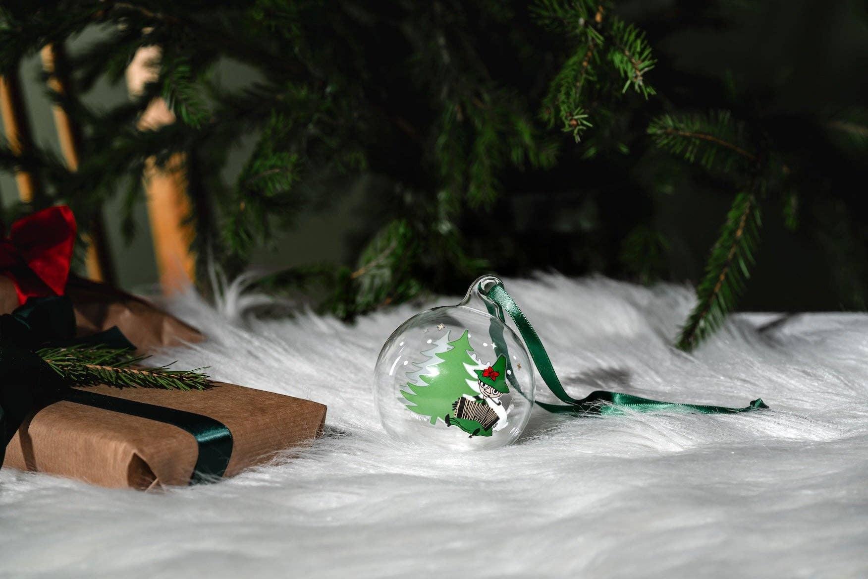 Moomin glass Christmas bauble with green tree design and ribbon, under fir tree on faux fur