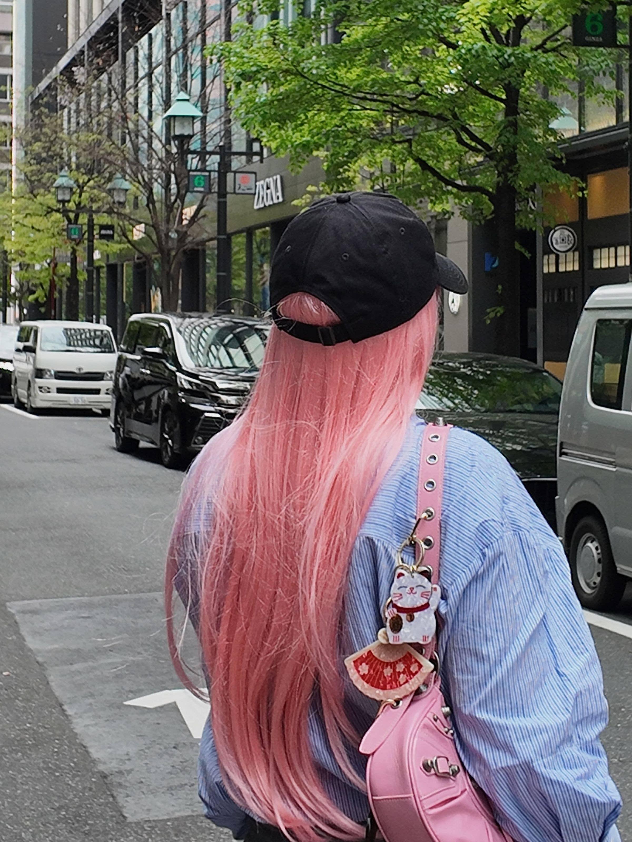 Person with long pink hair, black cap, blue shirt, and pink leather bag with lucky cat charm in city street