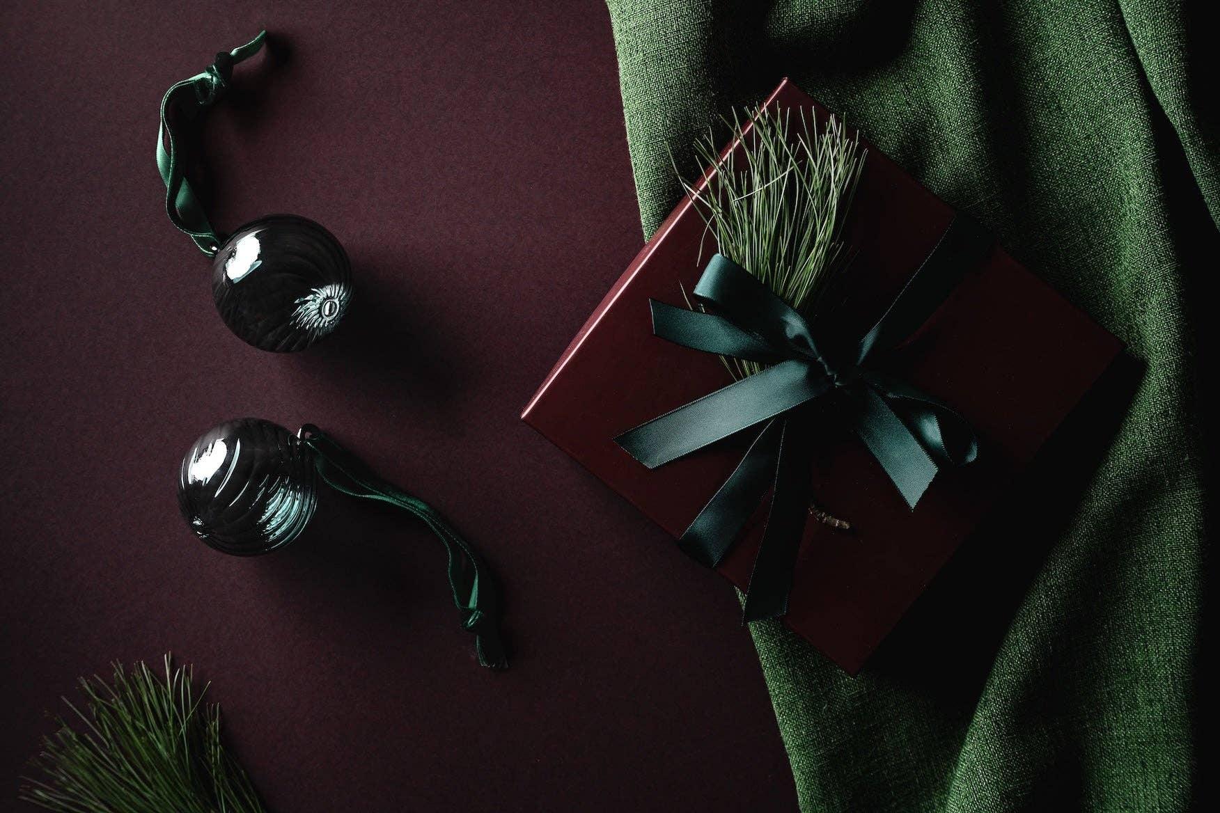 Elegant burgundy gift box with dark green ribbon, glass ornaments, pine, on green fabric