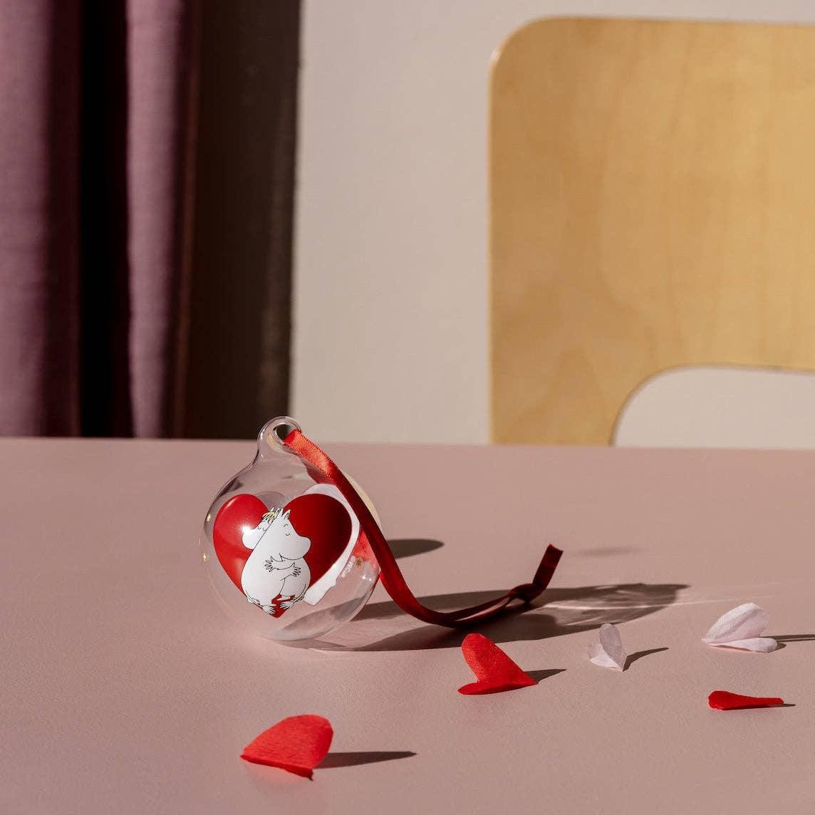 Moomin glass ornament with red heart and ribbon on pastel pink table, premium decor.