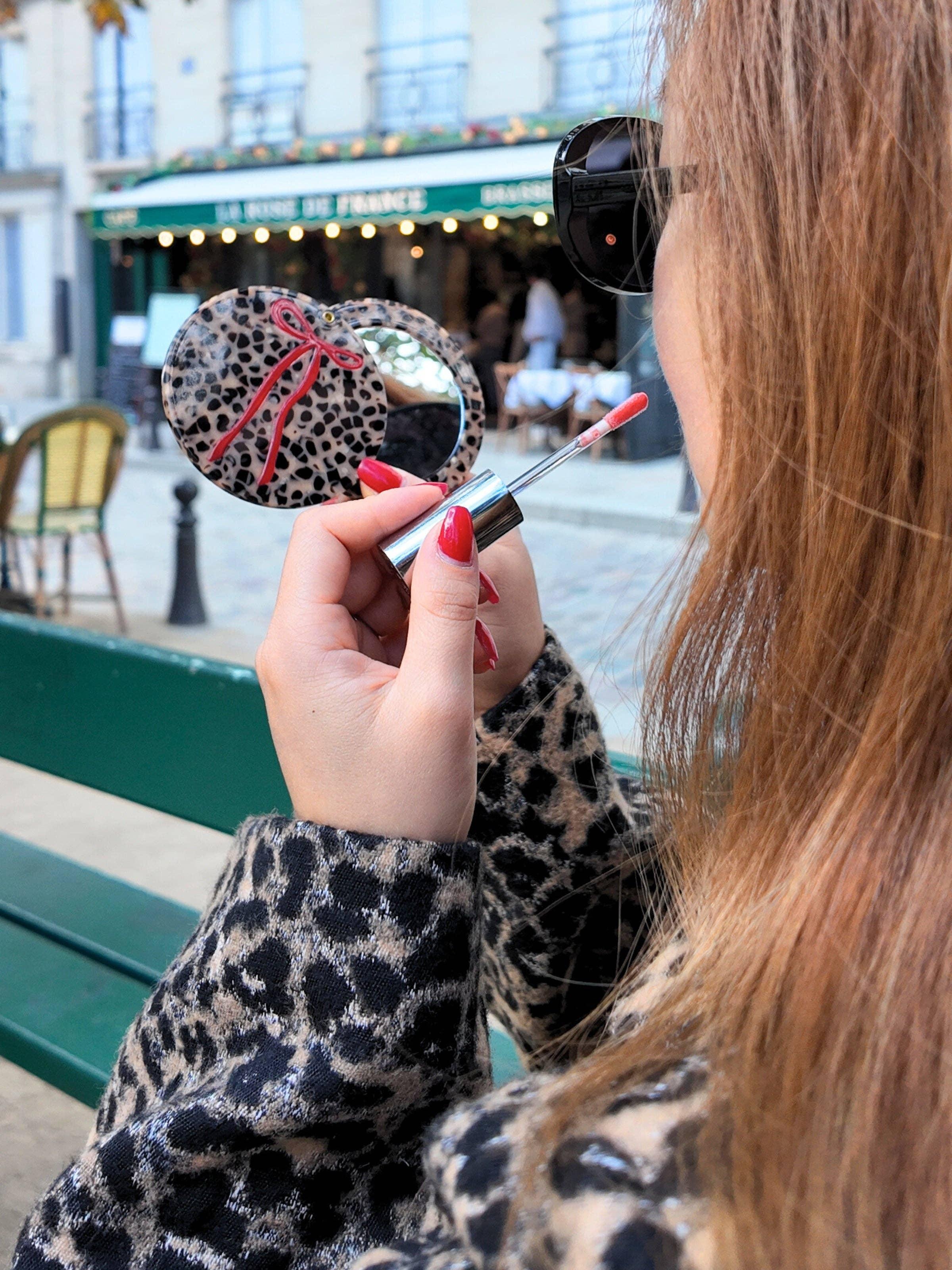 Balmain leopard print acrylic pocket mirror with red ribbon, woman applying lip gloss outdoors.