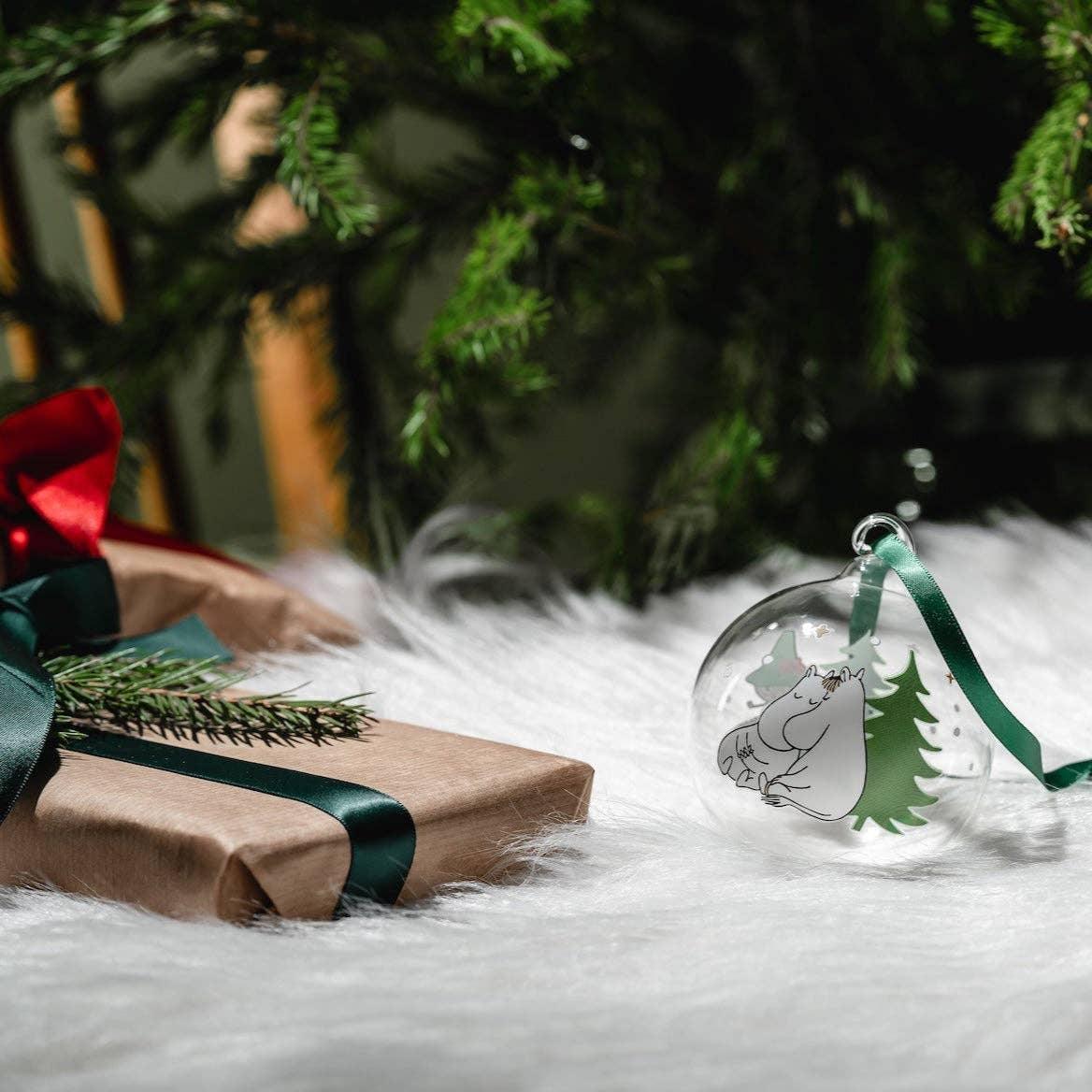 Moomin glass Christmas bauble with green tree design, on white fur under a decorated tree