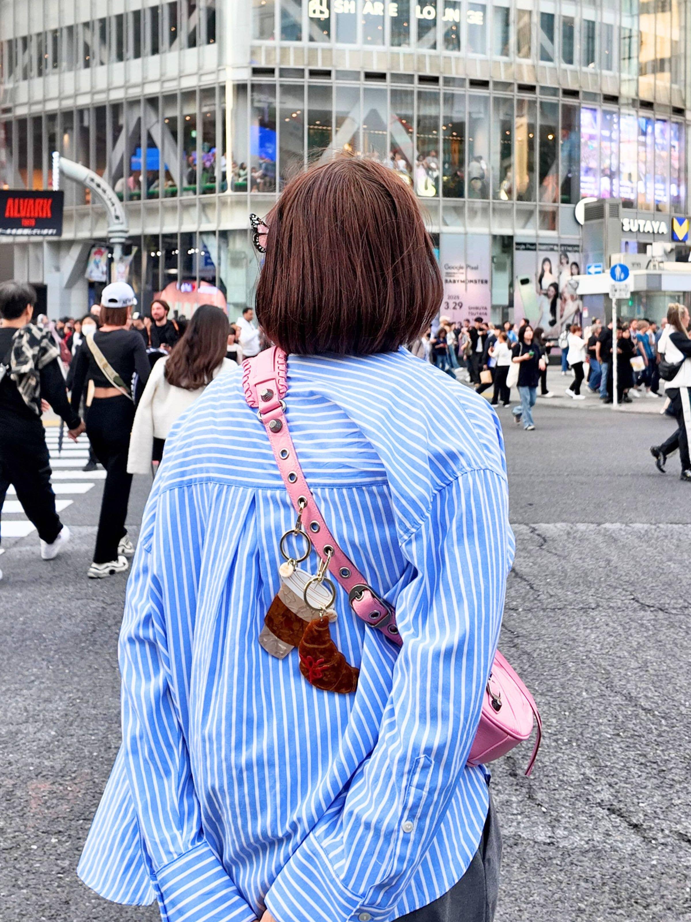 Person wearing blue striped shirt and pink leather bag with acrylic keychains, urban street scene