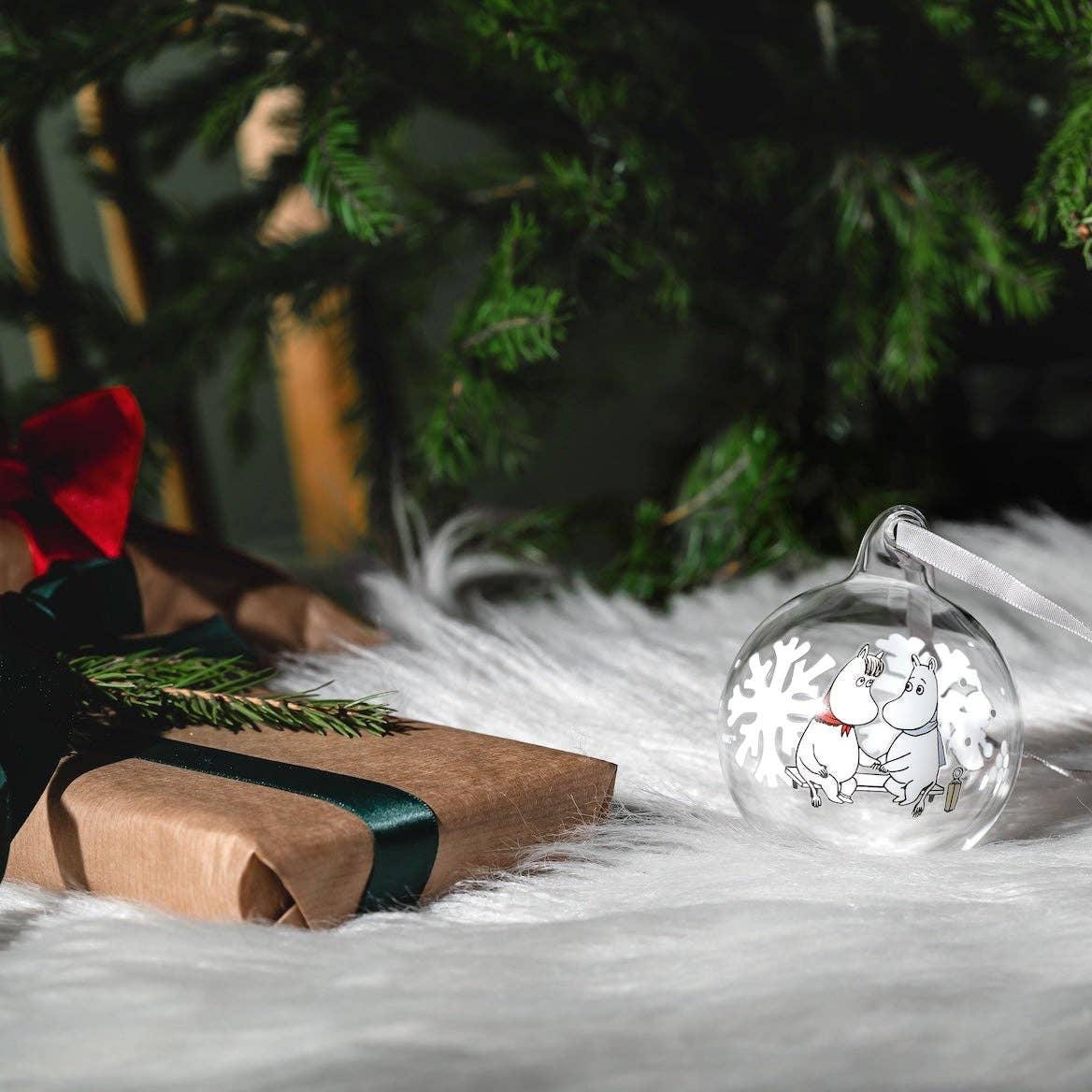 Moomin glass Christmas bauble with snowflake, clear and white, on fur under tree by gift