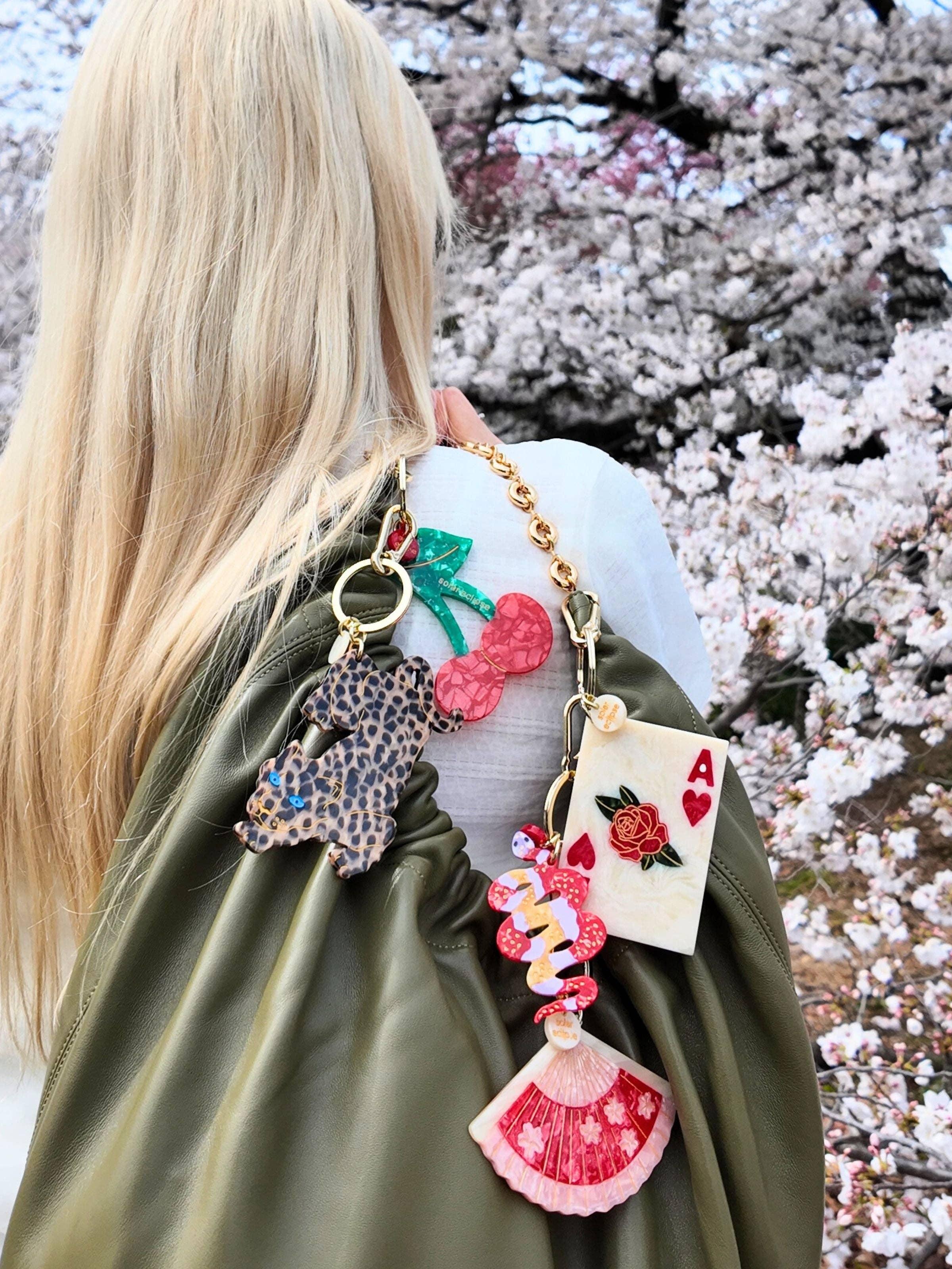 Roxanne Assoulin olive leather bag with colorful resin charms, gold chain, cherry blossoms background
